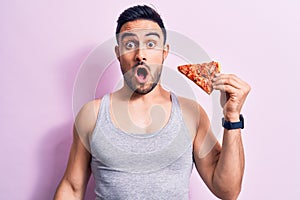 Young handsome man with beard eating slice of Italian pizza over isolated pink background scared and amazed with open mouth for