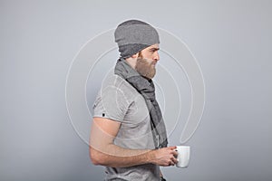 Young handsome man with the beard in grey cap and scarf standing by the grey wall turned in profile holding cup of tea or coffe in