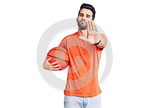 Young handsome man with beard hoilding basketball ball with open hand doing stop sign with serious and confident expression,