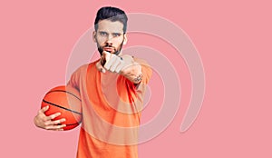 Young handsome man with beard hoilding basketball ball pointing with finger to the camera and to you, confident gesture looking