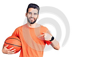 Young handsome man with beard hoilding basketball ball pointing finger to one self smiling happy and proud