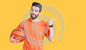 Young handsome man with beard hoilding basketball ball pointing thumb up to the side smiling happy with open mouth