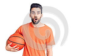 Young handsome man with beard hoilding basketball ball scared and amazed with open mouth for surprise, disbelief face