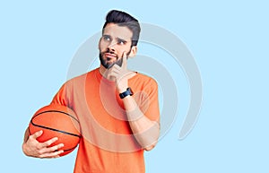 Young handsome man with beard hoilding basketball ball serious face thinking about question with hand on chin, thoughtful about