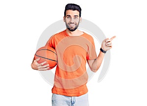 Young handsome man with beard hoilding basketball ball smiling happy pointing with hand and finger to the side