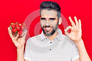 Young handsome man with beard holding bowl with strawberries doing ok sign with fingers, smiling friendly gesturing excellent