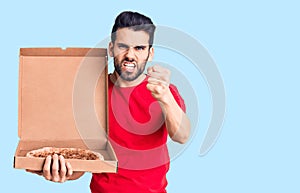 Young handsome man with beard holding delivery cardoboard with italian pizza annoyed and frustrated shouting with anger, yelling