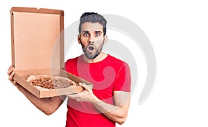 Young handsome man with beard holding delivery cardoboard with italian pizza scared and amazed with open mouth for surprise,