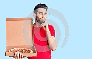 Young handsome man with beard holding delivery cardoboard with italian pizza serious face thinking about question with hand on