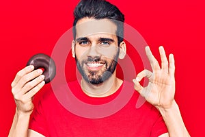 Young handsome man with beard holding donut doing ok sign with fingers, smiling friendly gesturing excellent symbol