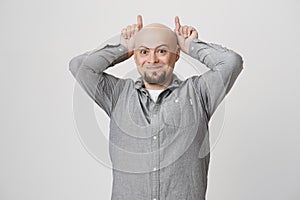 Young handsome man with a beard holding his hands above head making devil horns and smiling over white background. Or