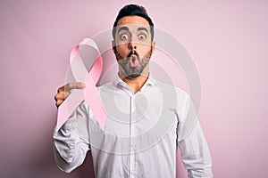 Young handsome man with beard holding pink cancer ribbon over isolated background scared in shock with a surprise face, afraid and