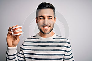 Young handsome man with beard holding plastic denture teeth over white background with a happy and cool smile on face