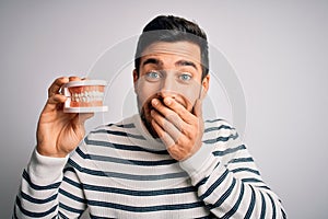 Young handsome man with beard holding plastic denture teeth over white background laughing and embarrassed giggle covering mouth