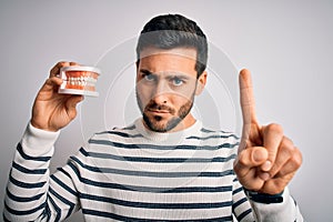Young handsome man with beard holding plastic denture teeth over white background Pointing with finger up and angry expression,