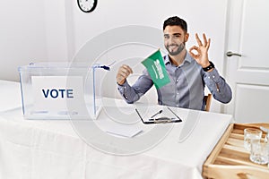 Young handsome man with beard at political campaign election holding arabia saudita flag doing ok sign with fingers, smiling photo