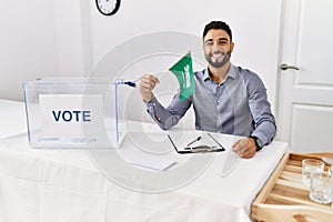 Young handsome man with beard at political campaign election holding arabia saudita flag looking positive and happy standing and photo