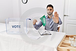 Young handsome man with beard at political campaign election holding arabia saudita flag with open hand doing stop sign with photo