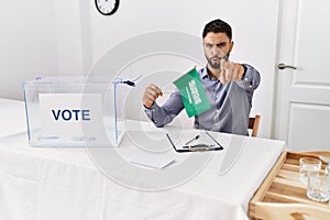 Young handsome man with beard at political campaign election holding arabia saudita flag pointing with finger to the camera and to photo