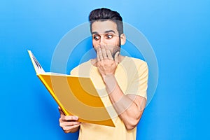 Young handsome man with beard reading book covering mouth with hand, shocked and afraid for mistake
