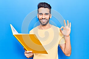 Young handsome man with beard reading book doing ok sign with fingers, smiling friendly gesturing excellent symbol