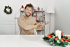 Young handsome man with beard sitting on the table by christmas decoration inviting to enter smiling natural with open hand