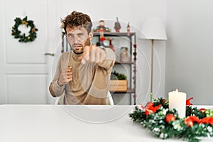 Young handsome man with beard sitting on the table by christmas decoration punching fist to fight, aggressive and angry attack,