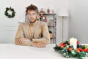 Young handsome man with beard sitting on the table by christmas decoration relaxed with serious expression on face