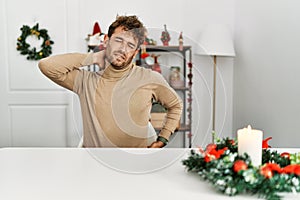 Young handsome man with beard sitting on the table by christmas decoration suffering of neck ache injury, touching neck with hand,