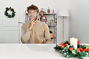 Young handsome man with beard sitting on the table by christmas decoration thinking worried about a question, concerned and