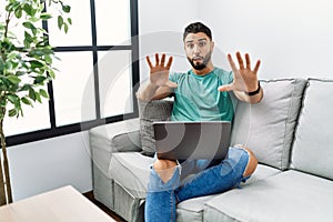 Young handsome man with beard using computer laptop sitting on the sofa at home afraid and terrified with fear expression stop