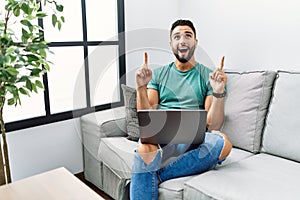 Young handsome man with beard using computer laptop sitting on the sofa at home amazed and surprised looking up and pointing with