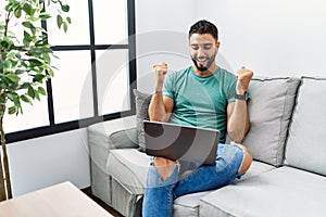 Young handsome man with beard using computer laptop sitting on the sofa at home celebrating surprised and amazed for success with