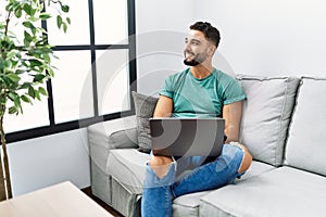 Young handsome man with beard using computer laptop sitting on the sofa at home looking away to side with smile on face, natural