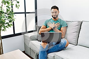 Young handsome man with beard using computer laptop sitting on the sofa at home smiling with hands on chest with closed eyes and