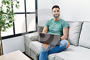 Young handsome man with beard using computer laptop sitting on the sofa at home smiling looking to the side and staring away