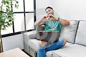 Young handsome man with beard using computer laptop sitting on the sofa at home smiling in love doing heart symbol shape with