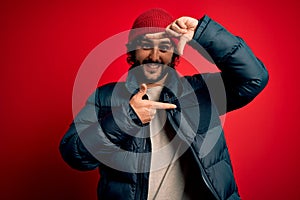 Young handsome man with beard wearing casual coat and wool cap over red background smiling making frame with hands and fingers