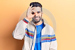 Young handsome man with beard wearing casual jacket doing ok gesture with hand smiling, eye looking through fingers with happy
