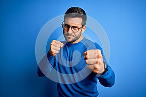 Young handsome man with beard wearing casual sweater and glasses over blue background Punching fist to fight, aggressive and angry