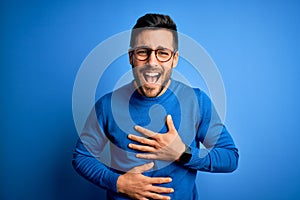 Young handsome man with beard wearing casual sweater and glasses over blue background smiling and laughing hard out loud because