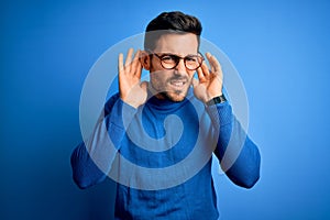 Young handsome man with beard wearing casual sweater and glasses over blue background Trying to hear both hands on ear gesture,