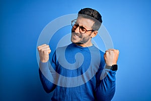 Young handsome man with beard wearing casual sweater and glasses over blue background very happy and excited doing winner gesture