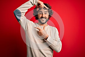 Young handsome man with beard wearing casual sweater standing over red background smiling making frame with hands and fingers with