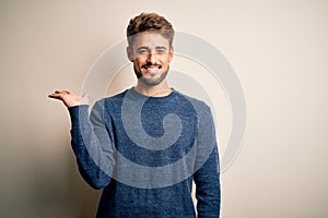 Young handsome man with beard wearing casual sweater standing over white background smiling cheerful presenting and pointing with