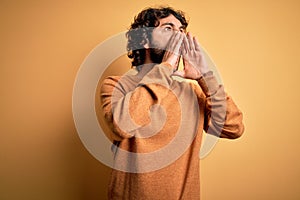 Young handsome man with beard wearing casual sweater standing over yellow background Shouting angry out loud with hands over mouth