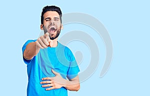 Young handsome man with beard wearing casual t-shirt laughing at you, pointing finger to the camera with hand over body, shame