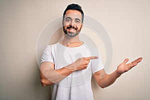 Young handsome man with beard wearing casual t-shirt standing over white background amazed and smiling to the camera while