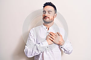 Young handsome man with beard wearing casual t-shirt standing over white background smiling with hands on chest, eyes closed with