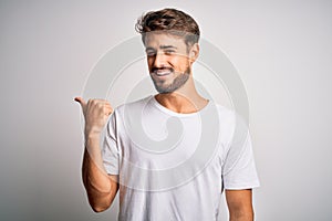 Young handsome man with beard wearing casual t-shirt standing over white background smiling with happy face looking and pointing